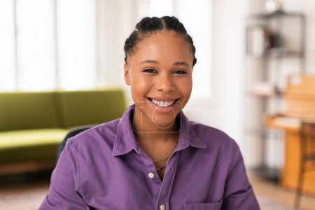 Téléchargez les photos : Portrait engageant d'une étudiante noire joyeuse portant une chemise violette, avec un sourire charmant regardant directement la caméra, assise dans une pièce lumineuse à la maison - en image libre de droit