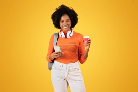 Photo for Smiling young black woman multitasking with headphones, holding a coffee cup and smartphone, dressed in an orange sweater and white pants against a yellow background, studio - Royalty Free Image