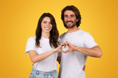 Photo for Affectionate european couple creating a heart shape with their hands, smiling joyfully, dressed in white shirts and blue jeans on a sunny yellow background, symbolizing love - Royalty Free Image