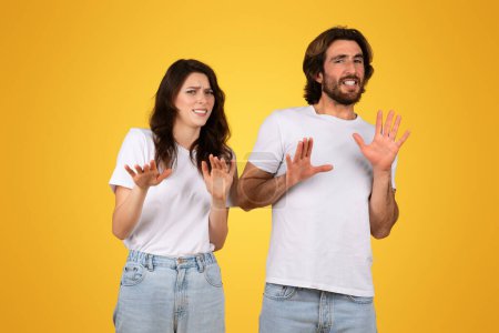 Téléchargez les photos : Un jeune couple européen inquiet gesticulant s'arrête avec les mains, montrant des signes de rejet ou de peur, avec des expressions anxieuses, vêtu de t-shirts blancs sur fond jaune - en image libre de droit