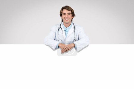 Young male doctor with stethoscope leaning over blank white banner, offering an engaging and helpful presence in healthcare setting, place for advertisement