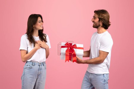 Photo for Happy caucasian couple exchanging gifts, with a man presenting a white box with a red ribbon to a delighted woman against a soft pink background, evoking feelings of joy and celebration - Royalty Free Image