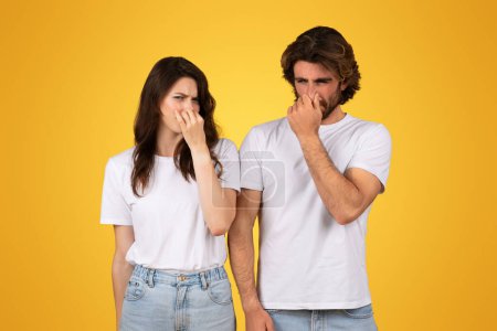 Photo for Distressed sad caucasian young couple in white shirts pinching their noses, displaying expressions of disgust, standing against a solid yellow background, studio. Bad stinks - Royalty Free Image