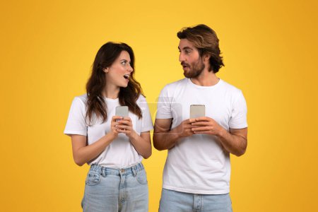 Téléchargez les photos : Engagé souriant couple caucasien millénaire tenant smartphones, la femme à l'air surpris et parlant à l'homme, à la fois en t-shirts blancs sur un fond jaune vif, studio - en image libre de droit