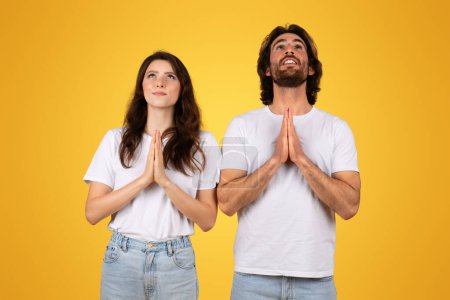 Photo for Hopeful european couple with palms pressed together in prayer, looking upwards with contemplative expressions, wearing white shirts and jeans against a mustard yellow background - Royalty Free Image