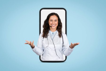 Friendly european female doctor with headset in giant smartphone screen gesturing and smiling at camera, having online appointment, blue background