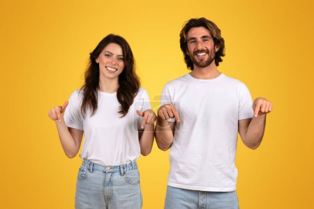 Téléchargez les photos : Femme européenne souriante et homme en chemises blanches pointant vers le bas avec les deux mains, semblant gaie et confiante, debout sur fond jaune, indiquant potentiellement un produit ou un texte ci-dessous - en image libre de droit