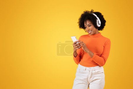 Photo for Engaged young African American woman with curly hair using smartphone and enjoying music through white headphones, wearing an orange sweater, on a yellow background, studio - Royalty Free Image