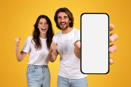 Photo for Exuberant caucasian young couple celebrating success with a woman clenching her fist and a man holding out a smartphone with a blank screen, on a yellow background, studio - Royalty Free Image