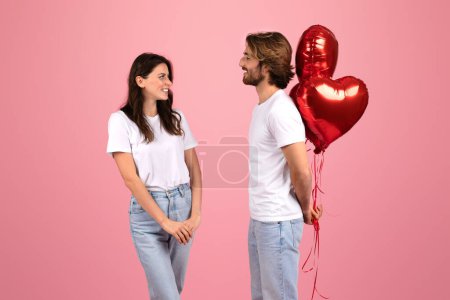 Téléchargez les photos : Un couple caucasien heureux se tient face à face avec une expression ludique, tenant des ballons rouges brillants en forme de cœur, symbolisant l'amour et la célébration, sur un fond rose doux - en image libre de droit