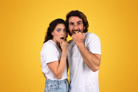 Photo for Shocked caucasian young couple in white t-shirts standing against a yellow background, with wide-eyed expressions of surprise, holding their hands to their mouths, studio - Royalty Free Image