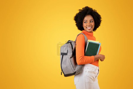 Photo for Confident young smart African American woman with a charming smile carrying a gray backpack and colorful notebooks, wearing a vibrant orange top on a yellow background, studio - Royalty Free Image