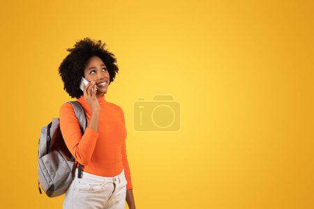 Foto de Radiante mujer afroamericana milenaria con cabello rizado natural usando un suéter naranja habla por teléfono, con una mochila gris sobre su hombro, contra un soleado telón de fondo amarillo - Imagen libre de derechos