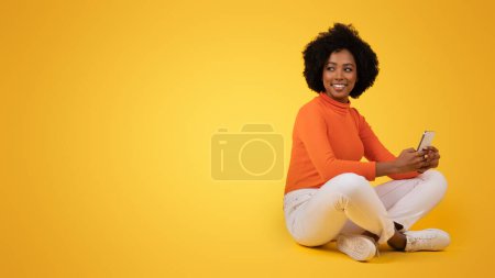 Foto de Mujer afroamericana milenaria con el pelo rizado sentado con las piernas cruzadas, sosteniendo un teléfono inteligente, con un cuello alto naranja y pantalones blancos, con una postura relajada sobre un fondo amarillo - Imagen libre de derechos