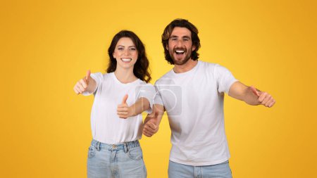 Téléchargez les photos : Couple européen radieux donnant les pouces levés, affichant approbation et bonheur, vêtus de tee-shirts blancs et de denim, avec des expressions joyeuses sur un fond jaune joyeux, studio - en image libre de droit