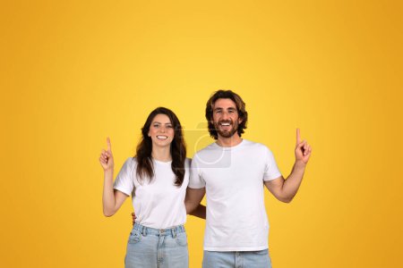 Téléchargez les photos : Homme et femme européens heureux et confiants pointant vers le haut, montrant de la positivité et s'amusant, à la fois en chemises blanches et en jeans en denim, avec un fond jaune vif - en image libre de droit