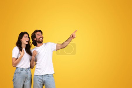 Foto de Feliz pareja caucásica extática con los ojos bien abiertos, hombre señalando y mirando hacia un lado con emoción, mujer animando, tanto en camisas blancas sobre un fondo amarillo, estudio - Imagen libre de derechos