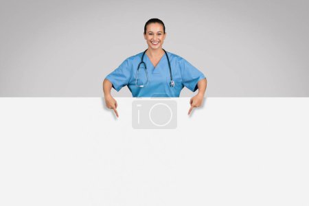Foto de Enfermera alegre en uniforme médico azul apuntando hacia abajo en el tablero blanco grande del cartel contra fondo gris, proporcionando la dirección clara y promoviendo el anuncio médico - Imagen libre de derechos