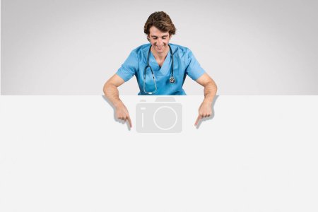 Foto de El alegre enfermero de uniforme azul se inclina hacia adelante sobre una pancarta blanca en blanco, sonriendo y apuntando hacia abajo en el espacio libre, perfecto para anuncios o mensajes - Imagen libre de derechos