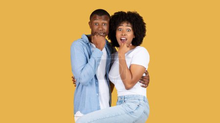 Photo for Excited frightened black young woman being lifted in a playful manner by a cheerful man, both showing surprised expressions, against a warm yellow background, studio, panorama - Royalty Free Image