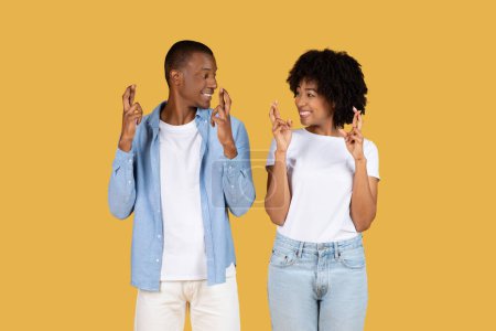 Photo for A joyful glad black young couple in casual wear happily making the fingers crossed gesture, looking at each other against a vibrant mustard yellow backdrop, studio. Wish, hope - Royalty Free Image