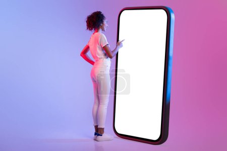 Foto de Vista trasera de una mujer usando un teléfono inteligente gigante, que simboliza el compromiso móvil a gran escala en el fondo de neón de degradado rosa a azul, espacio para publicidad - Imagen libre de derechos