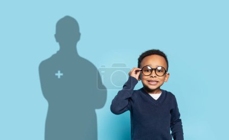 Téléchargez les photos : Un jeune garçon confiant avec des lunettes ajuste ses montures, souriant sur un fond bleu, son ombre projetant l'image d'un professionnel de la santé, symbolisant l'ambition et les objectifs futurs - en image libre de droit