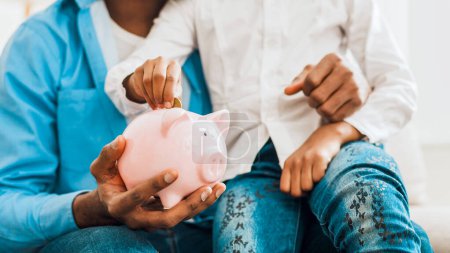 Photo for Girl and father putting coin into piggy bank, sitting on sofa at home - Royalty Free Image