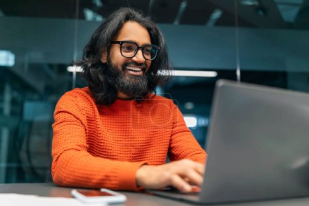 Téléchargez les photos : Positif travailleur barbu aux cheveux longs millénariste homme indien portant des lunettes IT professionnel assis au bureau au bureau sombre, travaillant sur ordinateur portable, travaillant sur le projet, l'espace de copie - en image libre de droit