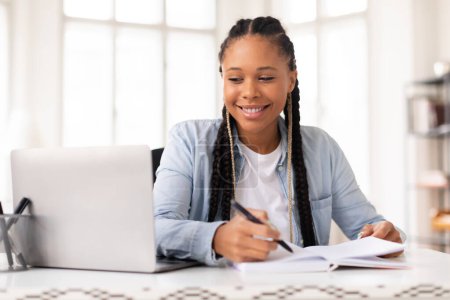 Foto de Sonriente joven estudiante negra con trenzas escribiendo notas, estudiando con computadora portátil en casa, teniendo una lección en línea mientras está sentada en una habitación luminosa - Imagen libre de derechos
