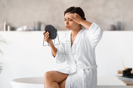 Foto de Mujer negra joven en albornoz blanco sosteniendo espejo, examinando críticamente su piel en la frente, mujer afroamericana preocupada notó cambios de edad o granos en la cara, sentada en el borde de la bañera moderna - Imagen libre de derechos