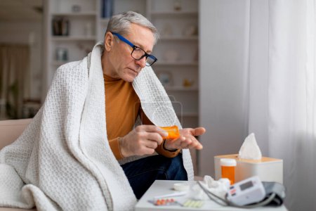 Foto de Abuelo enfermo con anteojos cubiertos de manta sentado en el sofá en casa, tomando pastillas, comprobando la presión arterial con tonómetro, anciano sintiéndose enfermo, espacio para copiar - Imagen libre de derechos