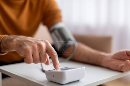 Photo for Cropped of senior man in casual outfit sitting on couch at home, checking blood pressure on his own. Pensioner using modern tonometer, pressing button on device, copy space - Royalty Free Image