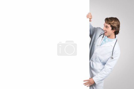 Alegre doctor masculino en bata blanca y estetoscopio sosteniendo y mirando una pizarra blanca grande en blanco, listo para consultas de pacientes o consejos de salud