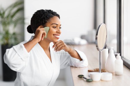 Téléchargez les photos : Femme noire souriante en peignoir blanc faisant routine de soins de la peau avec grattoir jade gua sha tout en étant assis devant le miroir dans la salle de bain, heureuse femme afro-américaine faisant massage facial, gros plan - en image libre de droit