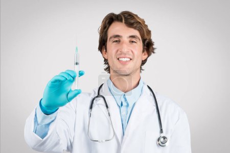 Foto de Alegre médico varón joven mostrando jeringa médica con aguja afilada, vestido con capa blanca con estetoscopio, sobre fondo gris limpio - Imagen libre de derechos