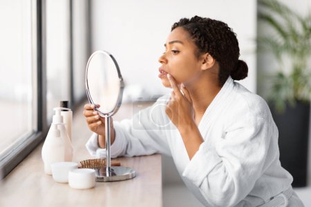 Téléchargez les photos : Inquiète jeune femme noire en peignoir blanc examinant l'épidémie d'herpès sur sa lèvre en regardant dans un miroir portatif, inquiète afro-américaine souffrant de problèmes de peau, gros plan - en image libre de droit
