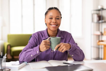 Foto de Feliz estudiante adolescente en camisa púrpura, sosteniendo la taza de café mientras se involucra con su computadora portátil y materiales de estudio, sonriendo a la cámara, sentado en el escritorio en casa - Imagen libre de derechos