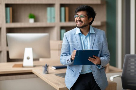 Photo for Cheerful Indian professional businessman holds clipboard and signing documents, celebrating successful business deals, standing in modern corporate office interior. Copy space - Royalty Free Image