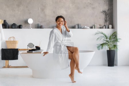 Foto de Sonriente mujer negra en albornoz blanco sentada en el borde de la lujosa bañera independiente en el interior del baño de diseño minimalista, feliz mujer afroamericana xuding relajación y elegancia - Imagen libre de derechos