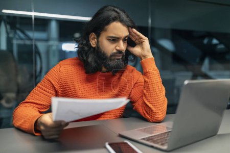 Photo for Stressed bearded millennial indian man entrepreneur sitting at dark office, holding papers financial report, looking at laptop computer screen, touching his head. Bankruptcy in business - Royalty Free Image