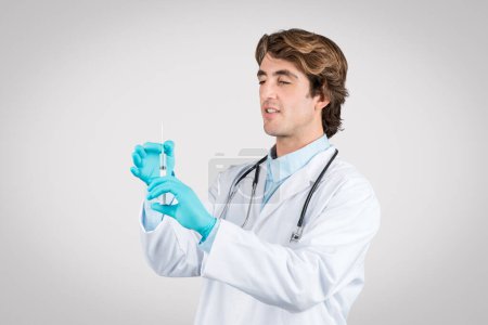 Foto de Médico masculino enfocado con estetoscopio examinando jeringa para vacunación, vestido con bata blanca y con guantes estériles azules sobre fondo gris - Imagen libre de derechos