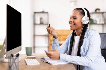 Photo for Smiling black lady with long braids wears stylish headphones, engaging in conversation while sitting at her computer in bright, modern workspace, studying at home - Royalty Free Image