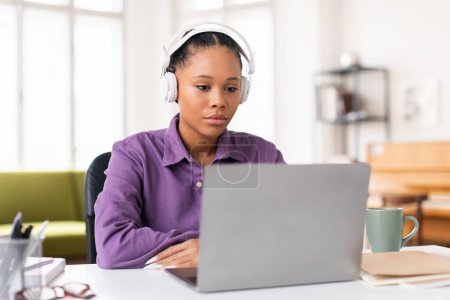 Téléchargez les photos : Sérieux afro-américaine adolescent dame à l'aide d'un ordinateur portable regardant l'écran tout en portant des écouteurs blancs dans l'environnement de la maison, montrant la concentration et la concentration - en image libre de droit