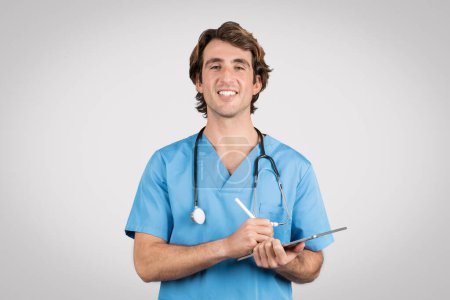 Confident male nurse in blue scrubs with stethoscope around his neck, smiling while writing on clipboard, representing professional medical care and patient record keeping