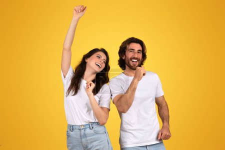 Photo for Exuberant european woman raising her fist in victory and joyous man laughing alongside, both enjoying a celebratory moment in white t-shirts, set against a yellow background - Royalty Free Image