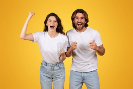 Téléchargez les photos : Jeune couple européen énergique aux poings levés célébrant une victoire, exsudant joie et excitation, portant des t-shirts et des jeans blancs sur un fond jaune vif, studio - en image libre de droit