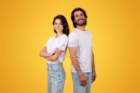 Photo for Side-by-side, a confident european millennial woman and a beaming man stand back-to-back in plain white t-shirts and light blue jeans against a sunny yellow backdrop, studio - Royalty Free Image