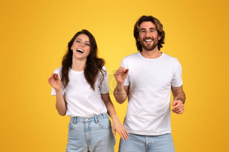 Téléchargez les photos : Jeune femme européenne rayonnante et homme en tee-shirts blancs riant et dansant, affichant un sentiment de liberté et de plaisir, parfaitement complété par le fond jaune énergique - en image libre de droit