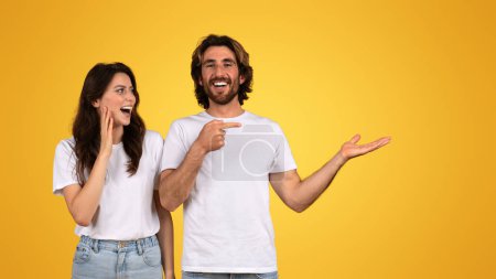 Téléchargez les photos : Joyeux couple européen interagissant et riant, avec l'homme pointant vers la femme comme si elle partageait une blague, tous deux portant des t-shirts blancs, profitant d'un moment léger sur un fond jaune - en image libre de droit